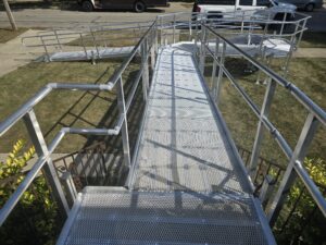 A metal wheelchair ramp with railings zigzags over a grassy lawn, providing accessible entry to a sidewalk and street where several vehicles are parked.