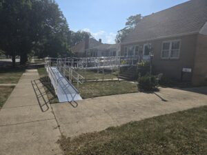 A metal wheelchair ramp leads from the sidewalk to the front door of a brick house, crossing the lawn and connecting to the porch steps. The house is on a sunny street with trees and other houses nearby.