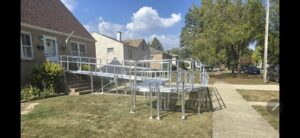 A metal wheelchair ramp with railings curves from a homes front door to the sidewalk, crossing a grassy yard on a sunny day. Nearby houses, trees, and a blue sky with scattered clouds are visible.