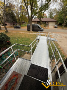 A metal wheelchair ramp leads from a house to the sidewalk, surrounded by autumn leaves. A black van is parked on the street, and trees with fall foliage are in the background. The word ACCESS is in the lower right corner.