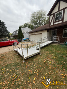A metal wheelchair ramp with railings leads from a grassy lawn to the front door of a house with a brick and siding exterior. The ramp passes by a raised garden bed. ACCESS company logo appears in the bottom right corner.