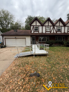 A metal wheelchair ramp with handrails leads from a driveway to the entrance of a two-story house with brown trim. Fallen leaves cover the lawn, and the sky is overcast. The Access logo is in the lower right corner.