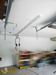 A modern garage ceiling with open garage door tracks, a garage door opener, and wiring hanging down. Shelves and tools are visible along the back wall, with part of the image blurred. Access logo in the corner.