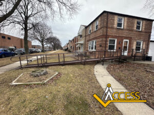 A long metal wheelchair ramp leads to the entrance of a brick two-story house on a corner lot. The yard has sparse grass and a tree stump. The ACCESS logo is in the bottom right corner.