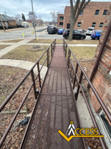 A metal wheelchair ramp with handrails extends from a brick building to a sidewalk. The ground is mostly bare with patches of grass. The Access logo is in the bottom right corner.
