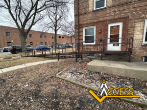 A metal wheelchair ramp leads from a concrete step to a sidewalk outside a brick building. Parked cars and leafless trees are visible in the background. An Access logo is in the lower right corner.