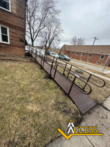 A brown metal wheelchair ramp with handrails leads from a sidewalk to the entrance of a brick building. Leafless trees and parked cars are visible in the background. The ACCESS logo is at the bottom right corner.