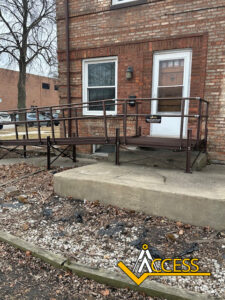 A brown metal wheelchair ramp leads to a white door at a brick buildings entrance. Bare trees and a leaf-covered ground surround the area. The word ACCESS with a yellow logo appears in the bottom right corner.