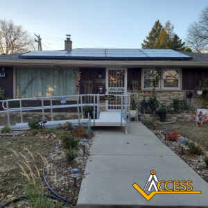 A single-story house with solar panels on the roof, a metal wheelchair ramp leading to the front door, and a concrete walkway through a landscaped yard; the ACCESS logo is in the lower right corner.