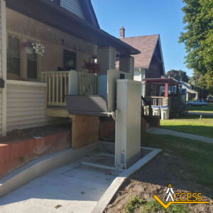 A wheelchair lift is installed at the front steps of a house, providing accessibility from the sidewalk to the porch. The Access Elevator & Lift logo is visible in the bottom right corner.