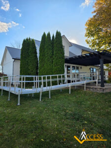 A white house with large green trees in front, featuring a metal wheelchair ramp leading to a covered patio. The Access logo is visible in the bottom right corner.