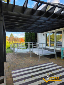 A metal wheelchair ramp leads from a brick patio under a black pergola to the entrance of a white house, with autumn trees visible in the background. The Access logo is in the lower right corner.