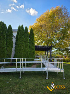A metal wheelchair ramp leads to the entrance of a building surrounded by green trees and grass. The sky is blue with a few clouds. The Access logo is in the lower-right corner.