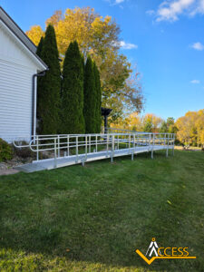 A long aluminum wheelchair ramp with handrails is installed alongside a white house, surrounded by green grass, trees, and a clear blue sky. The ACCESS logo is visible in the bottom right corner.