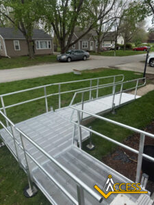 A metal wheelchair-accessible ramp with railings is installed on a grassy lawn, leading from a sidewalk to a house. Residential houses and parked cars are visible in the background. Access branding is in the corner.