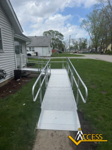 A metal wheelchair ramp with railings leads from the sidewalk to the entrance of a house. The yard is grassy, and other houses and trees are visible in the background. The Access logo is at the bottom right corner.