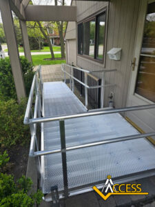 A metal wheelchair ramp with handrails leads up to the front door of a house. The walkway is bordered by bushes, and the word “ACCESS” with a logo appears in the lower right corner.