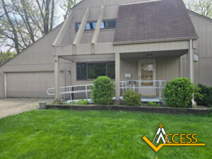 A beige house with a slanted roof has a ramp with railings leading to the front door. Bushes and green grass are in the yard. The logo “Access” is in the bottom right corner.
