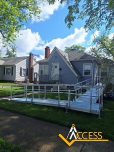 A small gray house with a white door and windows has a metal accessibility ramp leading from the sidewalk to the front entrance. Green grass and trees surround the home. ACCESS logo is in the lower right corner.