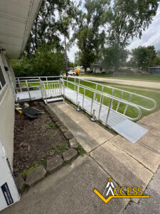 An aluminum wheelchair ramp with handrails is installed beside a house, providing accessible entry from a concrete path. Trees and houses are visible in the background. The ACCESS company logo is in the lower right corner.