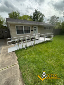 A beige house with a white door and windows has a metal wheelchair ramp leading from the front door to the sidewalk. The yard is green and the Access company logo is in the bottom right corner.