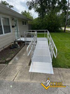 A metal wheelchair ramp with handrails leads from a concrete sidewalk to the entrance of a beige house. The ACCESS logo is in the bottom right corner of the image.
