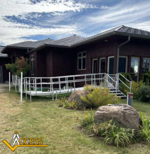 A brown house with large windows features a white metal wheelchair ramp leading to the entrance. The ramp is surrounded by green grass, shrubs, rocks, and a partly cloudy sky. The Access logo is in the bottom left corner.