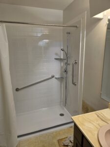 A bathroom with a walk-in shower featuring white subway tile, a showerhead with hose, grab bars, a curtain, and a small corner shelf. Part of a sink and countertop is visible in the foreground.