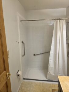 A bathroom with a walk-in shower featuring white subway tile, a glass door, a white shower curtain, and wall-mounted grab bars. The floor is light-colored, and a wooden door is partially open. A toilet paper holder is visible.