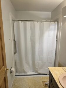A small bathroom with a closed white shower curtain, a silver grab bar on the wall, a toilet paper holder, a beige sink countertop, and a towel on the floor. The flooring is patterned vinyl.