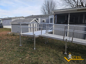 A metal wheelchair ramp with railings leads from the grassy yard up to the entrance of a gray manufactured home. The ramp is elevated and supported by metal posts. The Access brand logo appears in the bottom right corner.