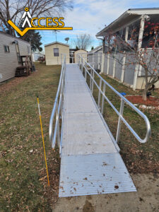 A long metal wheelchair ramp with handrails on both sides leads from a grassy lawn up to the entrance of a mobile home. The ramp is sturdy and accessible. A yellow pole stands near the bottom left side.