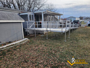 A metal wheelchair ramp with railings leads to the porch of a gray house, passing beside a shed. The ground is covered with grass and fallen leaves. The Access logo is in the bottom right corner.