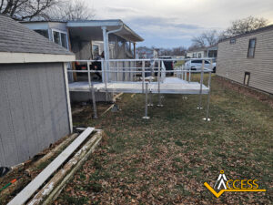 A metal wheelchair ramp with railings leads from a mobile homes porch to the yard, passing between a gray shed and another mobile home. Fallen leaves cover the ground and the sky is mostly cloudy. ACCESS logo in the corner.