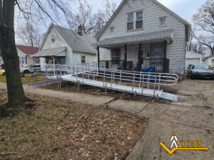 A metal wheelchair ramp leads from the sidewalk to the porch of a gray house with white trim. The yard is leaf-covered, and nearby houses and parked cars are visible. The Access logo is in the bottom right corner.