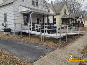 A metal wheelchair ramp with railings leads from the sidewalk to the porch of a white house. The yard has brown leaves, and there is a grill near the ramp. Several houses are visible in the background.