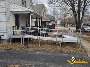 A metal wheelchair ramp with railings leads from a driveway up to the front porch of a house. The ramp is installed over grass and concrete. Nearby, other houses and parked cars are visible. ACCESS logo in the lower right corner.