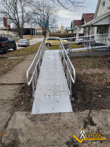 A metal wheelchair ramp with handrails extends from a sidewalk to a house entrance; it is surrounded by bare ground and some fallen leaves. The Access logo is visible in the bottom right corner.