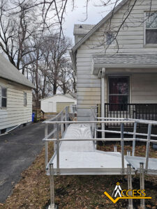 A metal wheelchair ramp leads from a driveway to the porch of a gray house, passing through a yard with bare trees. The ramp has railings on both sides. A small white shed is visible in the background.