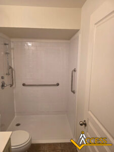 A bathroom with a walk-in shower featuring white subway tile, a grab bar on the back wall, a handheld showerhead, and a toilet next to the shower. The ACCESS logo is in the bottom right corner.