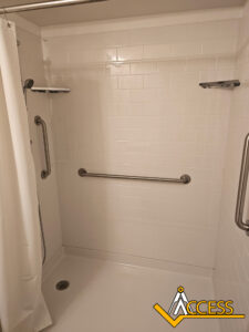 Accessible shower with white tile walls, a curtain, a handheld showerhead, two metal grab bars on the walls, corner shelves, and the ACCESS logo in the bottom right corner.