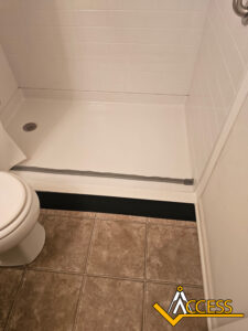 A walk-in shower with a low threshold and a horizontal gray safety bar, next to a toilet. The floor is tiled, and the Access company logo is at the bottom right corner.