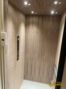 A modern elevator interior with wood-patterned walls, a control panel on the left, a handrail on the right, and ceiling spotlights. The Access company logo appears in the bottom right corner.