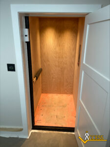 An open residential elevator with unfinished wooden walls and floor, illuminated by a ceiling light. A black handrail is mounted on the left wall. The Access logo is visible in the bottom right corner.