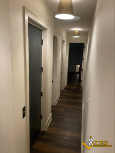A well-lit hallway with dark wooden floors, white walls, and several doors on the left side. Ceiling lights illuminate the space, and a round mirror is visible at the end of the hallway. ACCESS logo in bottom right corner.