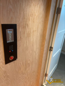 A wooden elevator panel with buttons and a digital floor display is mounted on a wall next to a partially open white door. The Access company logo is visible in the bottom right corner.