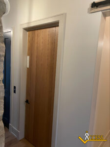 A wooden door with vertical panels set in a white frame is shown in a hallway with a stone wall on one side. There is a light switch on the wall and the Access logo in the lower right corner.