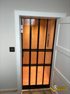 A home elevator with wooden interior walls and floor, black metal gate, and a control panel on the wall. The elevator door is open and the Access company logo is visible in the bottom right corner.