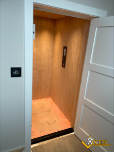 A residential elevator with wood-paneled walls, an open white door, and a control panel inside. The floor has protective plywood covering. The Access logo is visible in the bottom right corner.