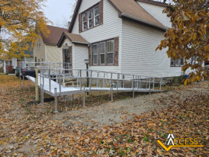 A beige house with brown roof has a silver metal wheelchair ramp leading from the front porch to the sidewalk. Fallen autumn leaves cover the yard. The ACCESS logo is in the bottom right corner.